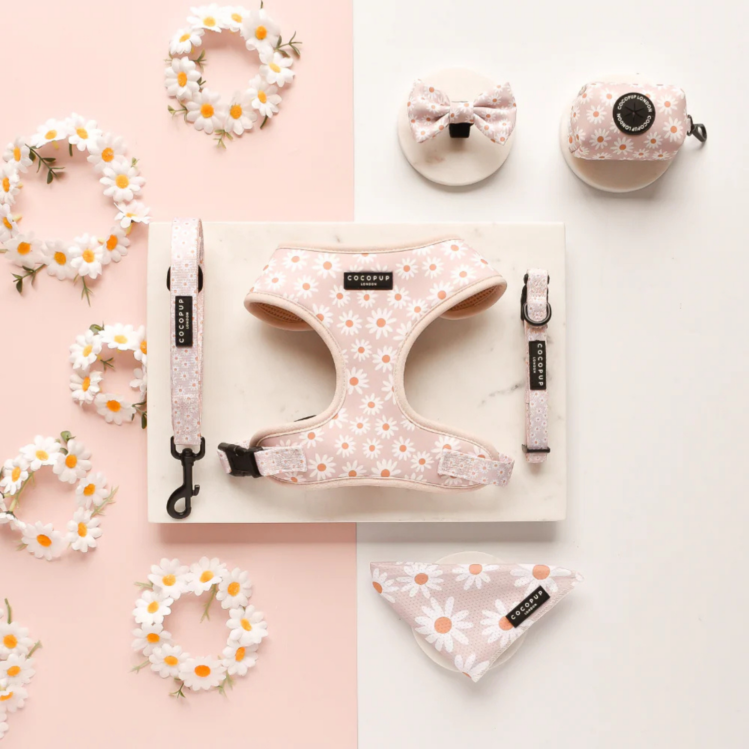 Pet accessories including a harness, collar, and bandana with floral patterns on a pink background.